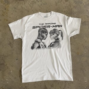 K&ouml;nnte beinhalten: Wei&szlig;es T-Shirt mit einem Schwarz-Wei&szlig;-Aufdruck von Spider-Man und einer Frau. Der Text "THE AMAZING SPIDER-MAN" ist in Schwarz &uuml;ber der Grafik aufgedruckt. Das T-Shirt ist aus weichem Material. Ein Klassiker f&uuml;r Superhelden-Fans.