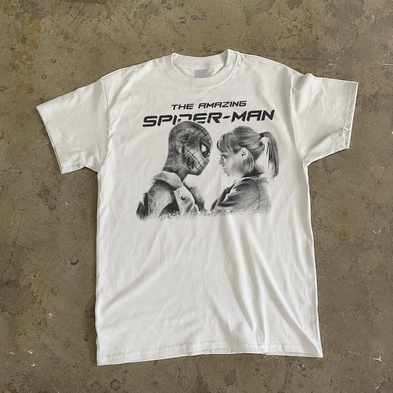K&ouml;nnte beinhalten: Wei&szlig;es T-Shirt mit einem Schwarz-Wei&szlig;-Aufdruck von Spider-Man und einer Frau. Der Text "THE AMAZING SPIDER-MAN" ist in Schwarz &uuml;ber der Grafik aufgedruckt. Das T-Shirt ist aus weichem Material. Ein Klassiker f&uuml;r Superhelden-Fans.