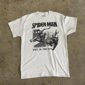 Puede incluir: Camiseta blanca con un gr&aacute;fico en blanco y negro de Spider-Man. El texto "SPIDER-MAN" est&aacute; encima de la imagen y "ACROSS THE SPIDER-VERSE" debajo. La camiseta tiene cuello redondo y mangas cortas.