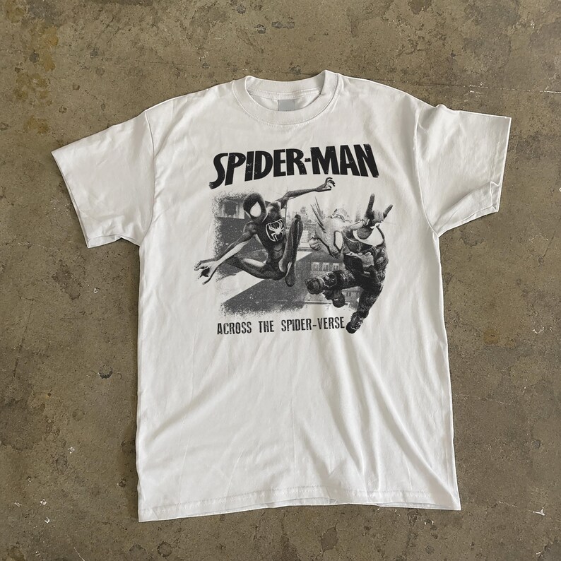 Puede incluir: Camiseta blanca con un gr&aacute;fico en blanco y negro de Spider-Man. El texto "SPIDER-MAN" est&aacute; encima de la imagen y "ACROSS THE SPIDER-VERSE" debajo. La camiseta tiene cuello redondo y mangas cortas.