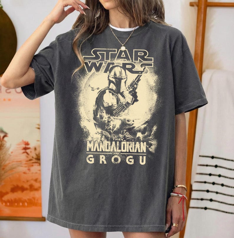 May include: Dark gray t-shirt featuring a graphic from the Star Wars series, "The Mandalorian." The design includes the show's logo, the Mandalorian character, and Grogu. The shirt has a vintage, worn look.
