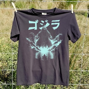 Könnte beinhalten: Ein anthrazitfarbenes Kurzarm-T-Shirt mit einem hellblauen Grafikdruck des Monsters Godzilla, das hinter einem kleinen Boot aus dem Wasser steigt, mit dem japanischen Text "ゴジラ" in Hellblau über dem Kopf der Kreatur gedruckt.