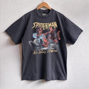 Może przedstawiać: Ciemnoszara koszulka z grafiką Spider-Mana. Projekt przedstawia czerwone i niebieskie postacie na tle miasta, z napisami "SPIDERMAN" i "No Way Home".
