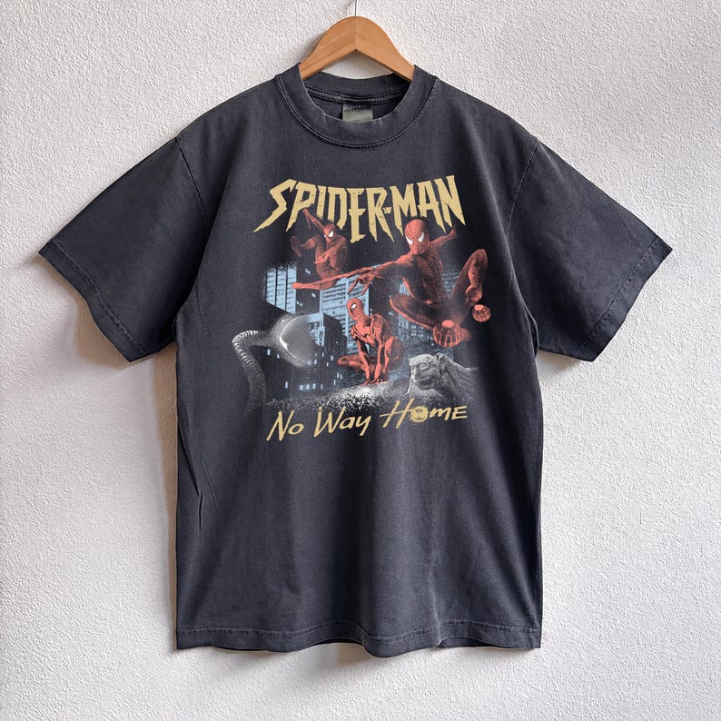 Może przedstawiać: Ciemnoszara koszulka z grafiką Spider-Mana. Projekt przedstawia czerwone i niebieskie postacie na tle miasta, z napisami "SPIDERMAN" i "No Way Home".