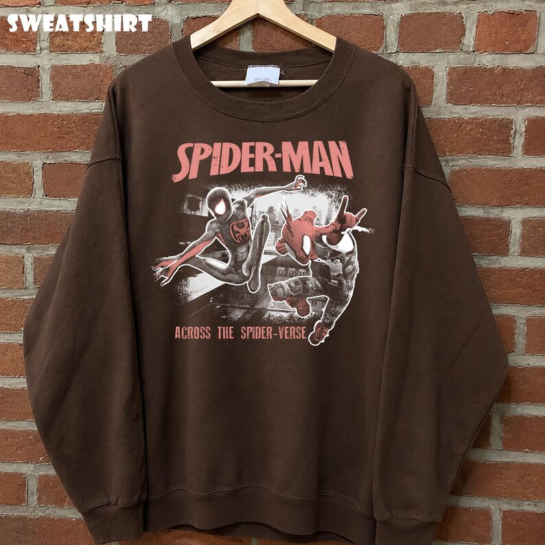Puede incluir: Sudadera marr&oacute;n con un gr&aacute;fico de Spider-Man. El texto "SPIDER-MAN" es rosa y "ACROSS THE SPIDER-VERSE" es blanco. Tiene cuello redondo y mangas largas. La palabra "SWEATSHIRT" est&aacute; en la parte superior.