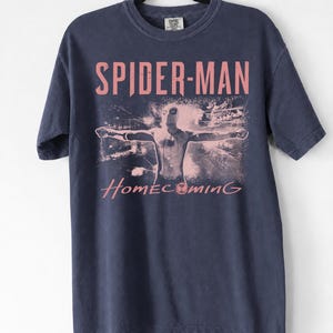 May include: Navy blue t-shirt featuring the words "SPIDER-MAN HOMECOMING" in pink lettering. The shirt displays an image of Spider-Man with outstretched arms. The t-shirt is hanging on a black hanger.