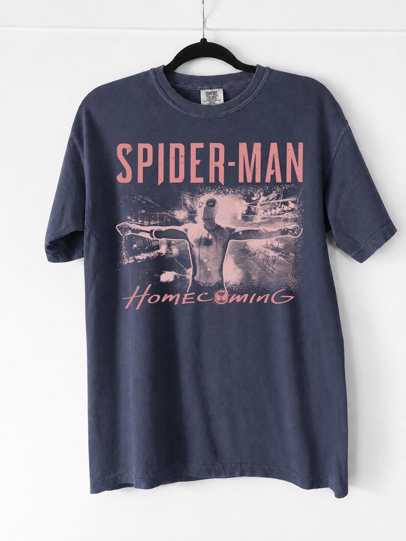 May include: Navy blue t-shirt featuring the words "SPIDER-MAN HOMECOMING" in pink lettering. The shirt displays an image of Spider-Man with outstretched arms. The t-shirt is hanging on a black hanger.