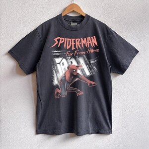 K&ouml;nnte beinhalten: Dunkelgraues T-Shirt mit dem Spider-Man-Logo und dem Text "Far From Home" in Rosa. Die Grafik zeigt Spider-Man in einem rot-schwarzen Anzug. Das T-Shirt h&auml;ngt an einem Holzb&uuml;gel.