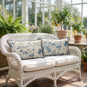May include: Two rectangular scatter cushions with a cream-coloured base and a blue floral toile pattern featuring birds and botanical elements. Each cushion is finished with a dark blue piped edge and is positioned on a white wicker loveseat.