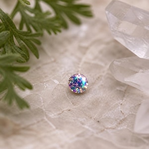 May include: A small, round, iridescent gemstone with a rainbow effect. The gem is set against a neutral, textured background, with a sprig of green foliage and clear quartz crystals in the background. The gem sparkles with colors of blue, purple, and green.