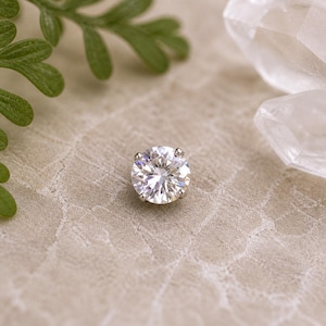 Op de afbeelding: Een sprankelende, rond geslepen diamant, gezet in een zilveren of platina klauwzetting. De diamant is het middelpunt, reflecteert licht en wordt omgeven door een neutrale achtergrond met groen en kristallen.