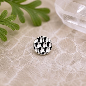 May include: A small, round, silver-toned button with a honeycomb pattern. The button has a textured surface with a series of interconnected, hexagonal shapes. The button is set against a neutral background, with a sprig of green leaves in the upper left corner.