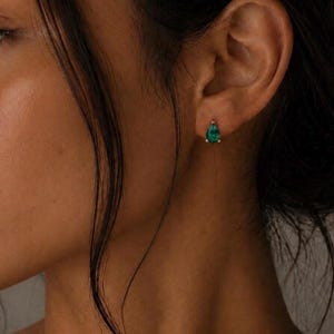 May include: A single teardrop-shaped earring featuring a vibrant green gemstone set in a metallic prong setting. The earring is positioned on the earlobe, showcasing a faceted surface that reflects light with a deep emerald hue and a polished finish.
