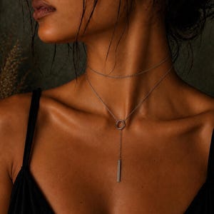 May include: A silver-toned lariat necklace featuring a delicate double-layered chain design. The necklace includes a circular ring connector that holds a thin, vertical rectangular bar pendant, creating a clean and simple drop-style aesthetic.