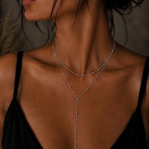 May include: A silver-toned layered necklace featuring two delicate chains adorned with small, evenly spaced metallic beads. The upper chain forms a circular collar, while the lower chain extends into a vertical drop, creating a Y-shaped silhouette.