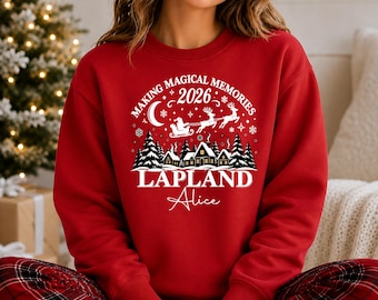 May include: A red crewneck sweatshirt featuring a white graphic print of a winter scene with houses, pine trees, a crescent moon, and Santa in a sleigh. The text reads Making Magical Memories 2026, Lapland, and the name Alice in script font.