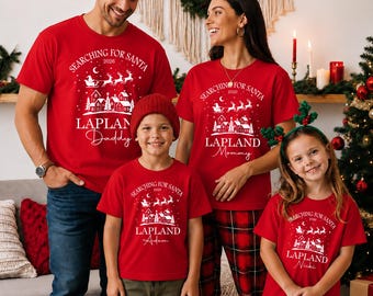 May include: A set of matching red short-sleeved t-shirts featuring a white graphic of a village, a sleigh with reindeer, and the text "SEARCHING FOR SANTA 2026 LAPLAND" with individual names "Daddy," "Mommy," "Adam," and "Nicki" printed on each shirt.