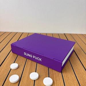 May include: A rectangular purple game board with the text SLING PUCK printed in white on the side. The game board features a hinged design and is accompanied by three small, round, white plastic pucks placed on the surface nearby.