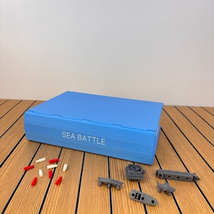 May include: A light blue rectangular board game case with the text SEA BATTLE printed in white on the front. Scattered in front of the case are several small red and white plastic pegs and four miniature gray plastic ship game pieces.