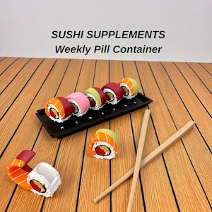 May include: A black rectangular weekly pill container labeled SUSHI SUPPLEMENTS Weekly Pill Container, featuring seven sushi-shaped compartments with white, orange, pink, green, and red details, accompanied by two light brown wooden chopsticks.