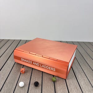 May include: A rectangular, metallic copper-coloured board game box with the text "SNAKES AND LADDERS" printed on the spine. The set includes one orange six-sided die, one white marble, one black marble, and one olive green marble.
