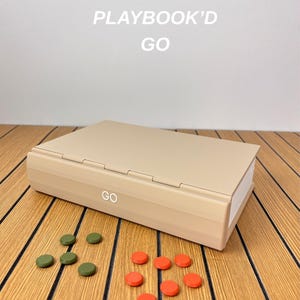 May include: A beige rectangular game case with the text PLAYBOOK'D GO printed above it and the word GO on the front edge. The case is accompanied by four small olive green circular tokens and six small bright orange circular tokens.