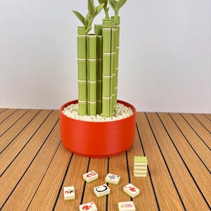 May include: A decorative bamboo plant sculpture in a round orange pot filled with white pebbles. Several small, square, light green tiles with various colorful graphic symbols and numbers are scattered in front of the pot, along with a small stack.