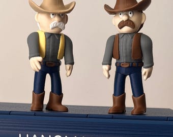 May include: Two cowboy figurines standing on a blue base labelled HANGMAN. The left figure wears a tan hat, yellow waistcoat, grey shirt, blue trousers, and brown boots. The right figure wears a brown hat, brown waistcoat, grey shirt, blue trousers, and brown boots.