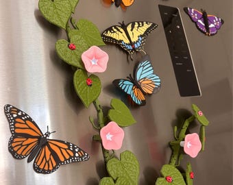 May include: A collection of decorative magnets featuring various butterfly species, including monarch and swallowtail, alongside a green vine with heart-shaped leaves, pink morning glory flowers, and small red ladybug accents.