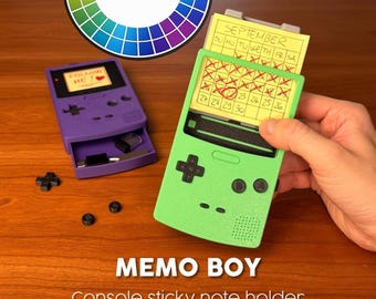 May include: A lime green and a purple handheld console-shaped sticky note holder. The green holder displays a yellow September calendar note. The purple holder features a drawer with small black buttons. Text reads: MEMO BOY Console sticky note holder.