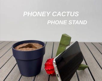 May include: A Phoney Cactus Phone Stand featuring a dark blue pot with brown soil, a red cactus, and a green cactus-shaped holder supporting a smartphone. The text PHONEY CACTUS PHONE STAND is visible above the objects in this desk accessory set.