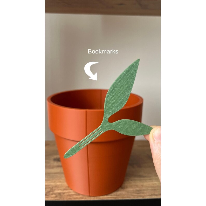 May include: A terracotta-colored plant pot with a green leaf-shaped bookmark. The bookmark is held near the pot, suggesting its use. The word "Bookmarks" is written above the pot.