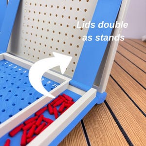 May include: A blue and white plastic board game with a grid featuring letters A through J and numbers 1 through 10. The game includes small red pegs and a blue lid that functions as a stand, with text stating Lids double as stands.