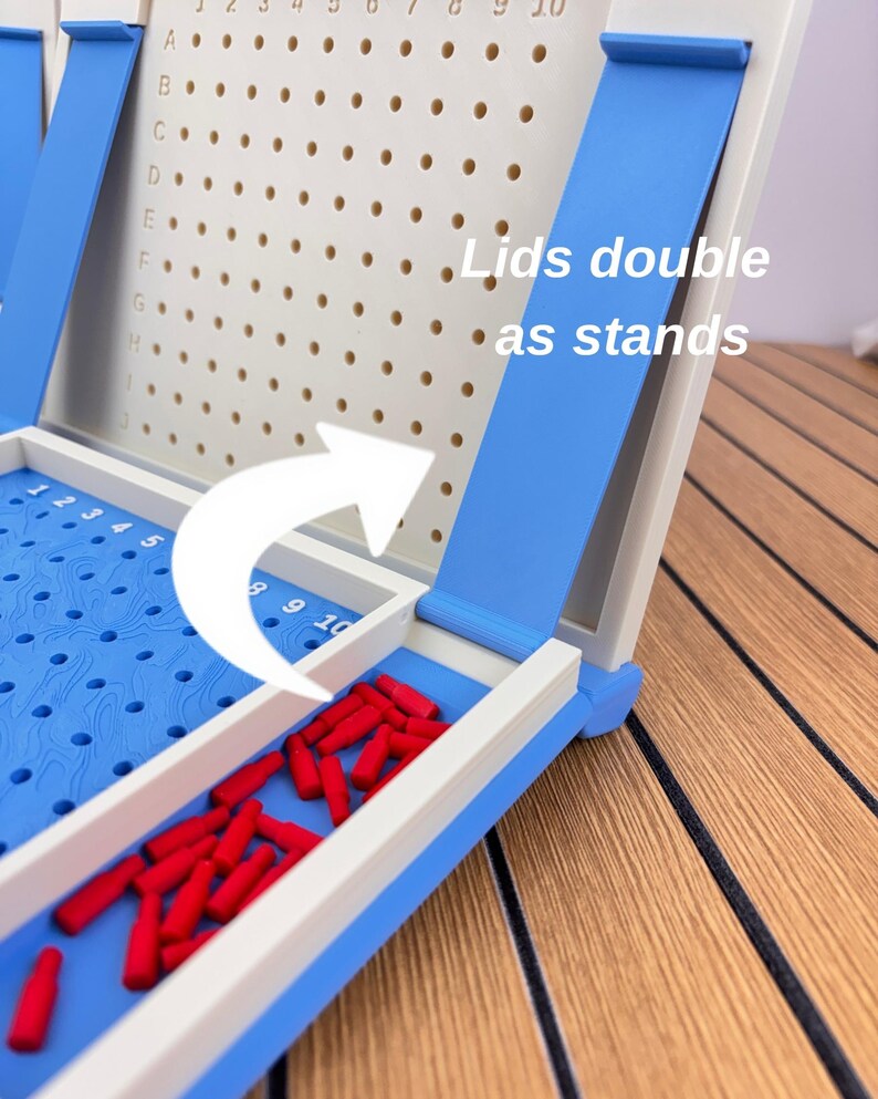 May include: A blue and white plastic board game with a grid featuring letters A through J and numbers 1 through 10. The game includes small red pegs and a blue lid that functions as a stand, with text stating Lids double as stands.