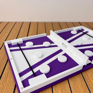 May include: A rectangular tabletop game board featuring a purple and white geometric pattern with white circular pucks and a black elastic cord stretched across the playing area. The board has white raised edges and a central divider.