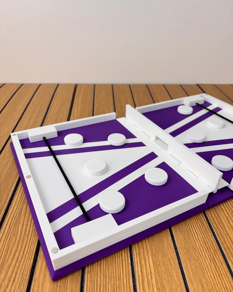 May include: A rectangular tabletop game board featuring a purple and white geometric pattern with white circular pucks and a black elastic cord stretched across the playing area. The board has white raised edges and a central divider.