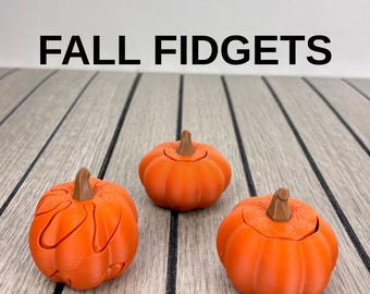 May include: Three orange pumpkin-shaped fidget toys with brown stems, arranged in a triangular formation. The text "FALL FIDGETS" is printed in bold black capital letters above the objects. Each pumpkin features a segmented, rounded design.