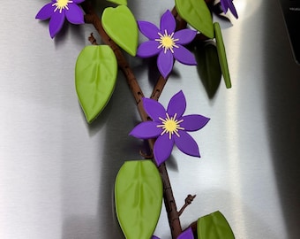 May include: A decorative vine magnet featuring a brown articulated stem with multiple bright green leaves and several purple flowers with yellow centres. The vine is arranged in a branching pattern with individual segments connected together.