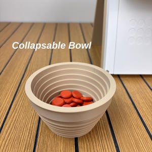 May include: A beige collapsible bowl with a ribbed, tiered exterior design. The interior of the bowl is filled with a collection of small, round, bright orange discs. The text "Collapsable Bowl" is visible in the upper left portion of the image.