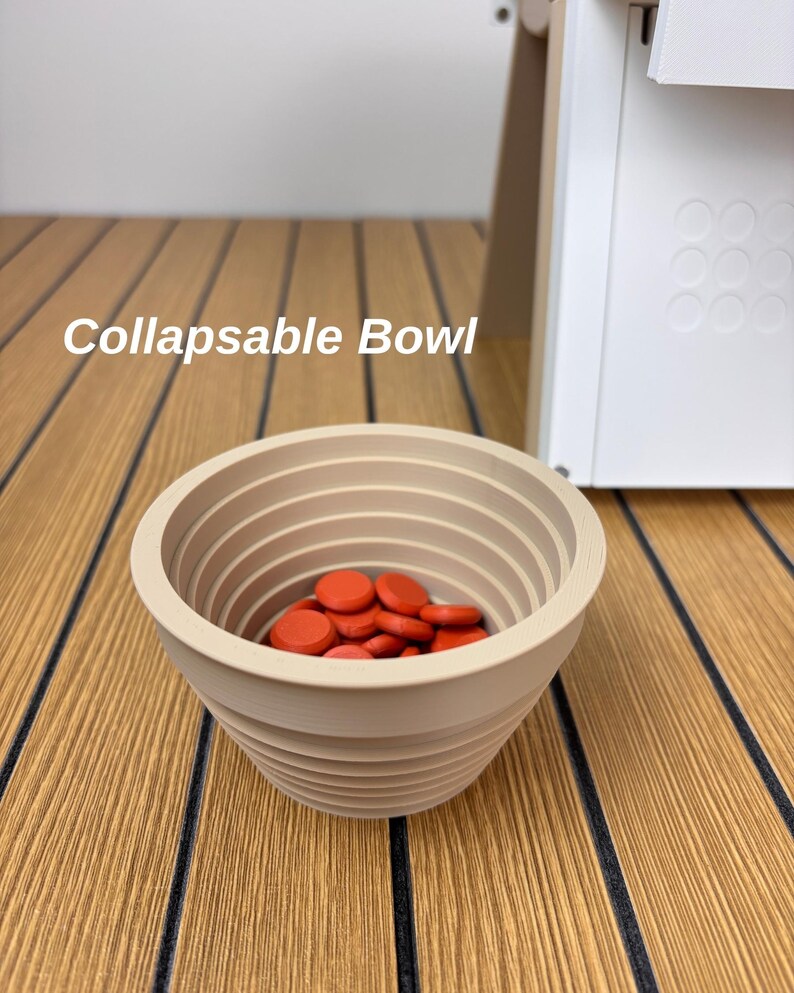 May include: A beige collapsible bowl with a ribbed, tiered exterior design. The interior of the bowl is filled with a collection of small, round, bright orange discs. The text "Collapsable Bowl" is visible in the upper left portion of the image.