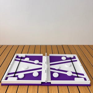 May include: A purple and white folding board game featuring a central divider, two elastic bands, and several circular white game pieces positioned on each side of the playing surface. The game board is constructed with a white frame and hinges.