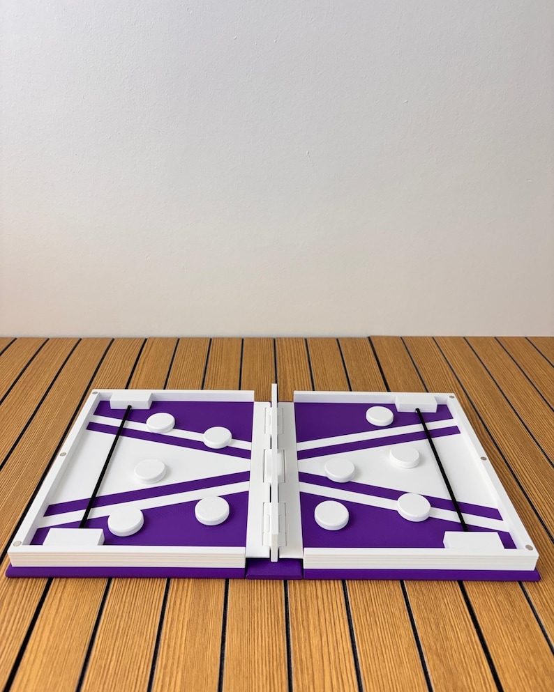 May include: A purple and white folding board game featuring a central divider, two elastic bands, and several circular white game pieces positioned on each side of the playing surface. The game board is constructed with a white frame and hinges.