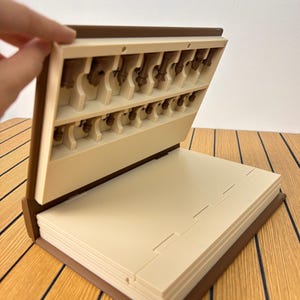K&ouml;nnte beinhalten: Ein braunes, buchf&ouml;rmiges Aufbewahrungsetui mit cremefarbenem Innenraum. Der obere Teil verf&uuml;gt &uuml;ber geformte Schlitze f&uuml;r kleine braune Schachfiguren, w&auml;hrend der untere Teil aus flachen, gestapelten cremefarbenen Platten besteht.