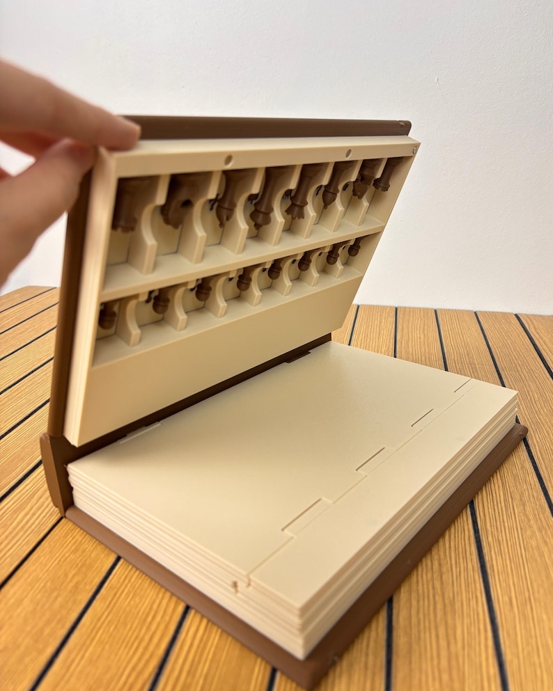 K&ouml;nnte beinhalten: Ein braunes, buchf&ouml;rmiges Aufbewahrungsetui mit cremefarbenem Innenraum. Der obere Teil verf&uuml;gt &uuml;ber geformte Schlitze f&uuml;r kleine braune Schachfiguren, w&auml;hrend der untere Teil aus flachen, gestapelten cremefarbenen Platten besteht.