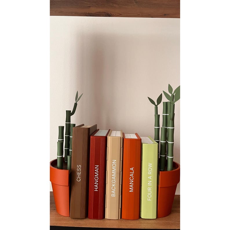 May include: A shelf arrangement featuring six books with game titles: Chess, Hangman, Backgammon, Mancala, and Four in a Row. The books are in various colors and are placed between two terracotta pots with bamboo plants.