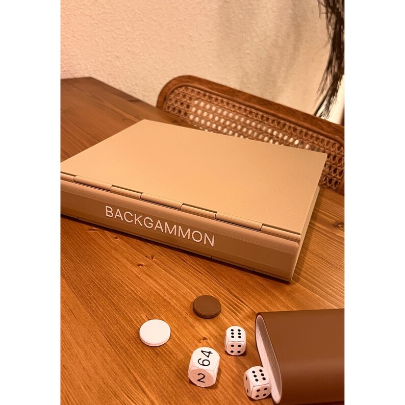 May include: A tan backgammon set with the word "BACKGAMMON" printed on the front. The set is closed and surrounded by game pieces, including dice and checkers. A brown leather dice cup is also present. The game is set on a wooden surface.