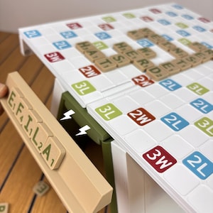 May include: A white word game board with a grid of colored squares labeled 2L, 3L, 2W, and 3W, featuring tan letter tiles spelling words. A tan tile rack holds tiles spelling E, F, I, L, A, with a green support piece attached to the board.