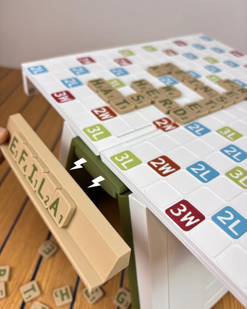 May include: A white word game board with a grid of colored squares labeled 2L, 3L, 2W, and 3W, featuring tan letter tiles spelling words. A tan tile rack holds tiles spelling E, F, I, L, A, with a green support piece attached to the board.