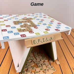 May include: A white and olive green folding board game with a grid surface featuring letter tiles spelling words like VEIN, HERD, and HAT. A tan tile rack displays the letters E, F, I, L, A, and a pile of loose letter tiles sits underneath.