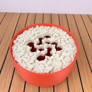 May include: A round, orange bowl filled with small, white, pebble-like objects. The bowl features a central, white, multi-holed insert that reveals a dark red interior, creating a contrast between the white surface and the deep red base.